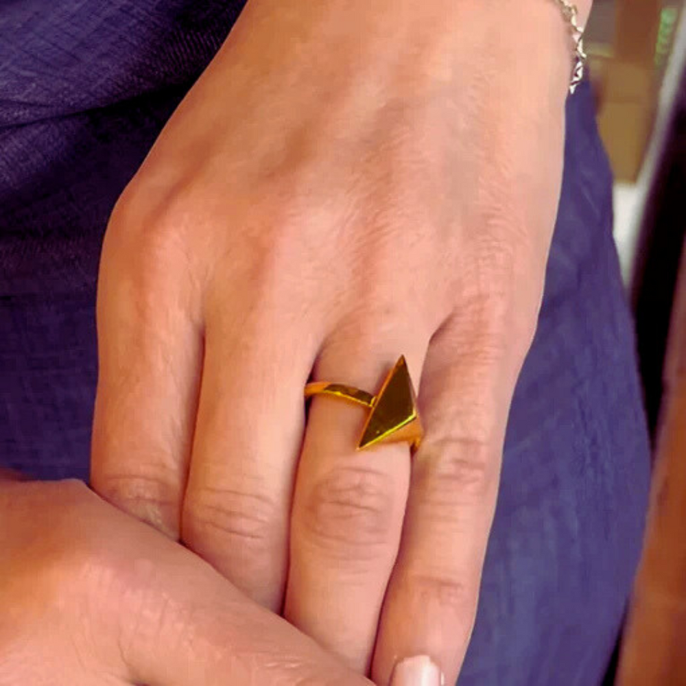 A hand wears the Atria Ring by Gallardo & Blaine Designs—a gold, geometric triangle ring—on the middle finger, resting gently on another hand.