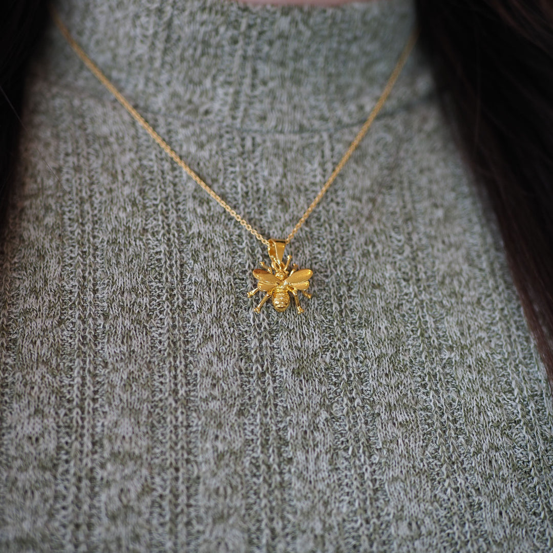 A close-up of a person in a gray-green ribbed sweater wearing the Gallardo & Blaine Designs Wildlife Pendant - Small Bee With Chain, a delicate gold bee-shaped necklace.