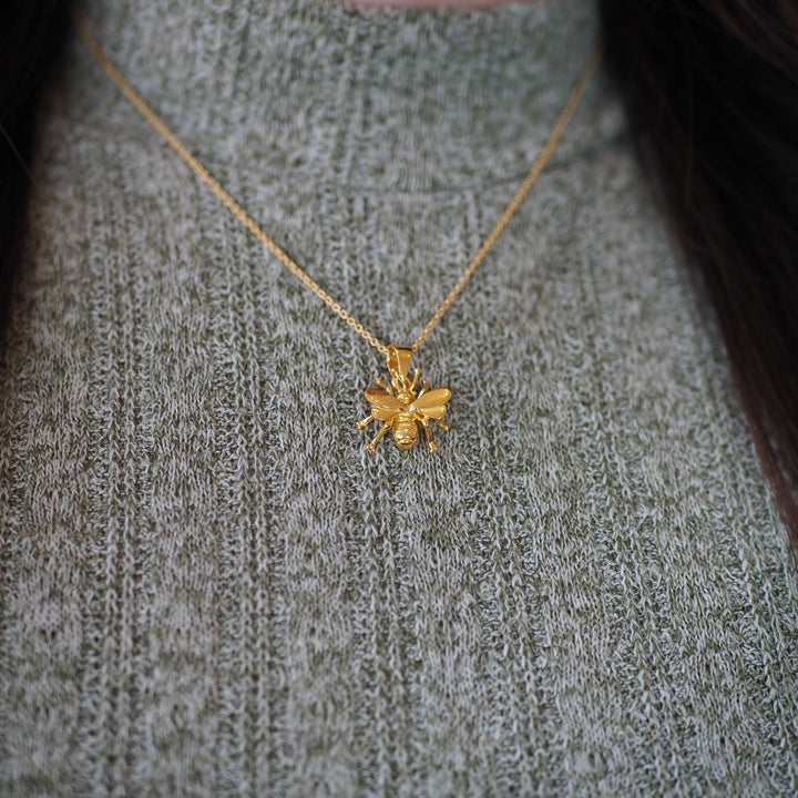 A close-up of a person in a gray-green ribbed sweater wearing the Gallardo & Blaine Designs Wildlife Pendant - Small Bee With Chain, a delicate gold bee-shaped necklace.