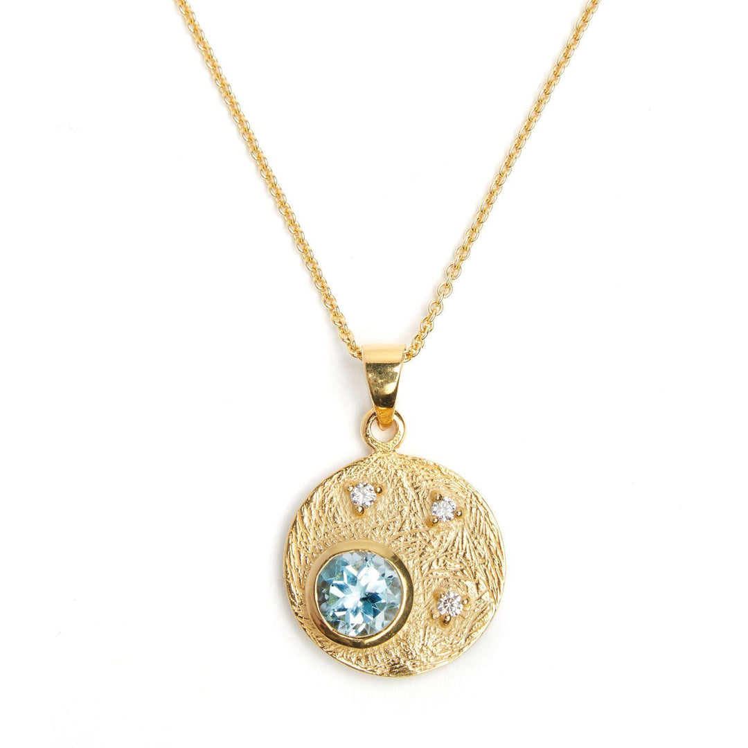 The Gallardo & Blaine Designs 9ct Yellow Gold Celeste Pendant - Blue Topaz & Natural Diamonds features a pendant in a textured circular design, adorned with a central blue topaz and three smaller white gemstones arranged symmetrically around it. The piece is completed by a thin, delicate gold vermeil chain.