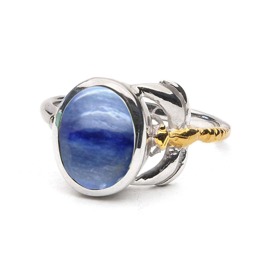 A silver ring featuring a large oval blue gemstone set in the center. The stone is held by intricate silver prongs with decorative yellow gold accents on either side of the band, creating an elegant and luxurious design. This timeless piece is perfect for adding a touch of sophistication to any outfit.
Introducing the Daydream Ring in various gemstones by Gallardo & Blaine Designs.