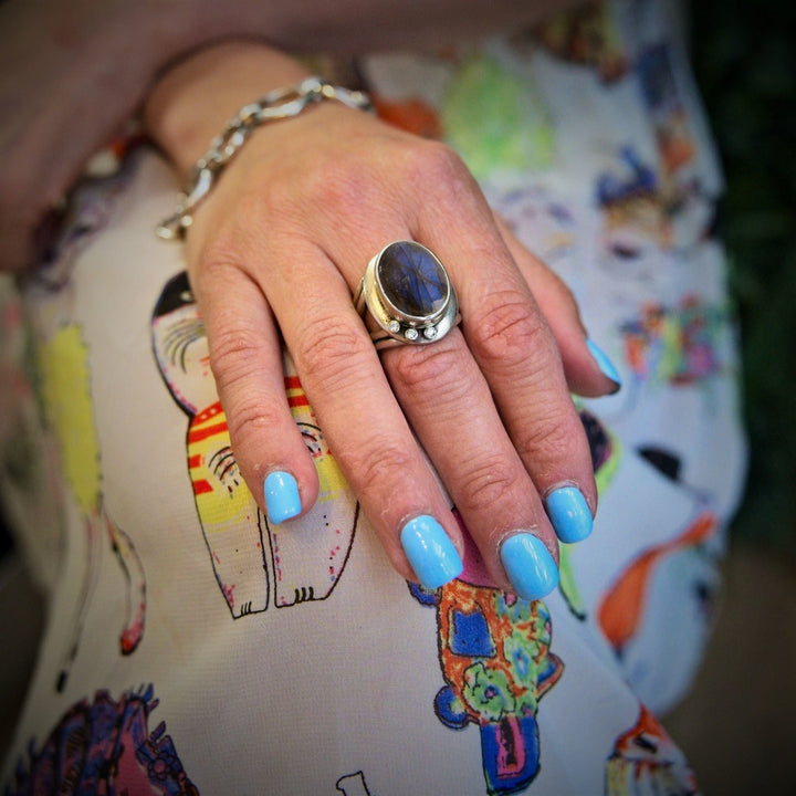A hand with light blue nail polish wears the Gallardo & Blaine Designs Eyetelia Ring in a gemstone and a sterling silver gold vermeil bracelet, resting on colorful fabric with cartoon cats.