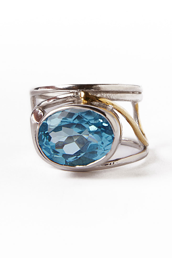 Gallardo & Blaine Designs' Horizontal Ring features an asymmetrical silver and gold band with a large oval-cut blue gemstone in the center, highlighted against a plain white background to showcase its intricate detail.