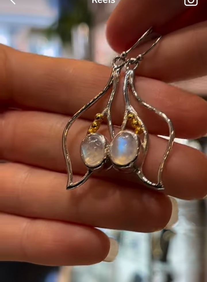 A hand holds Gallardo & Blaine Designs' Iris Earrings—silver and gold dangle earrings with opalescent oval gemstones and gold accents in an ornate, open metal design. The softly blurred background highlights their elegance.