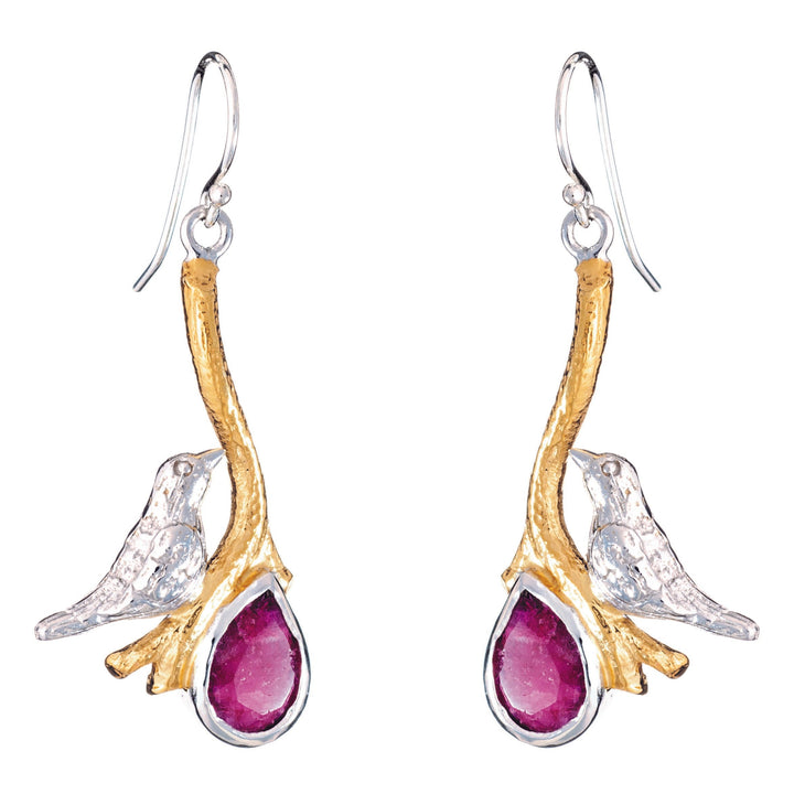 A pair of Lovebird Earrings in various gemstones from Gallardo & Blaine Designs features golden branches with silver birds perched on them, holding drop-shaped, deep red gemstones. The birds and branches are detailed with a shiny silver finish, while the gold adds a touch of elegance.