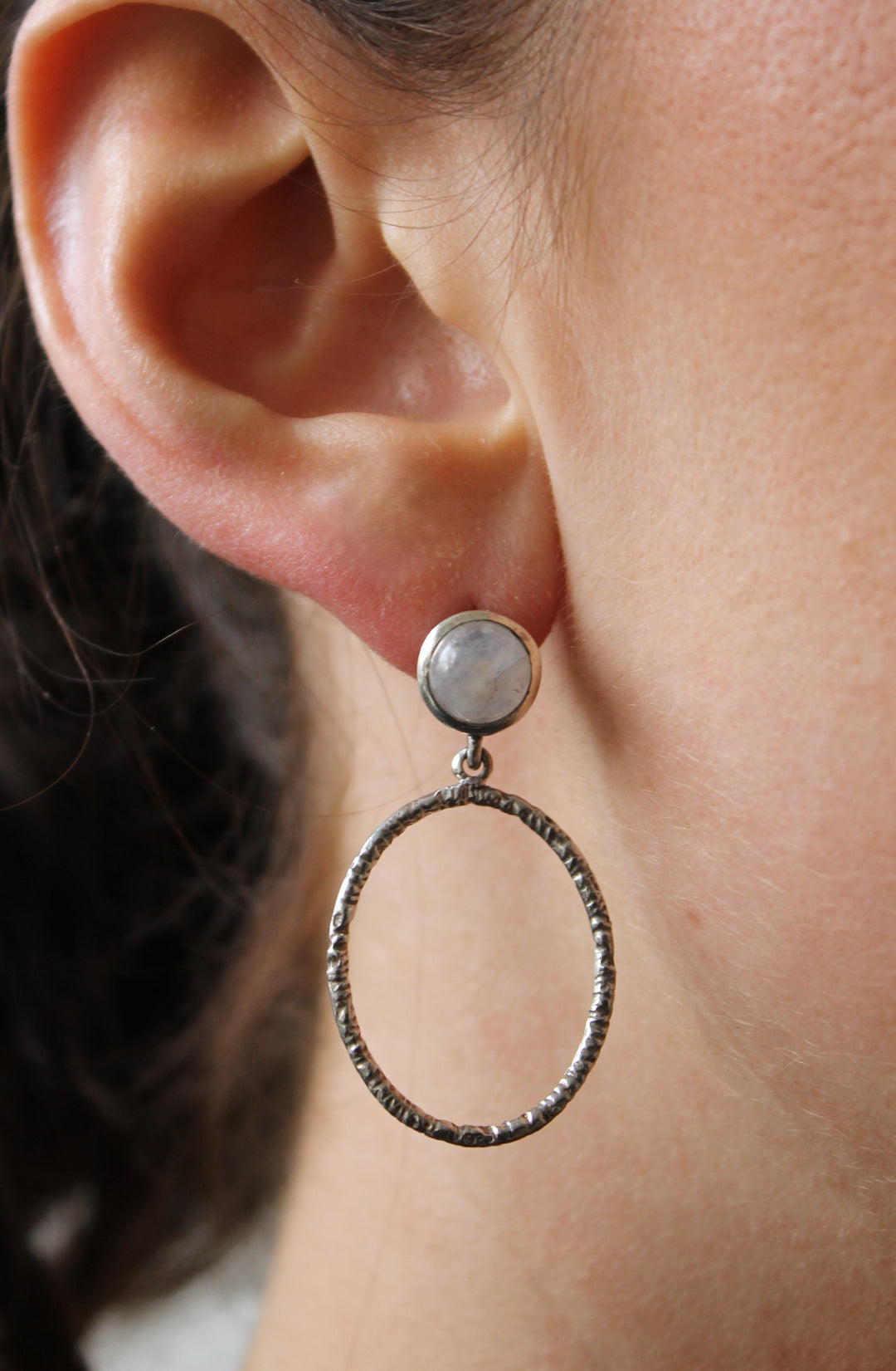 A close-up of a person’s ear wearing Gallardo & Blaine Designs’ Lunar Earrings, featuring various gemstones in a round stud with a large, textured circular hoop below.