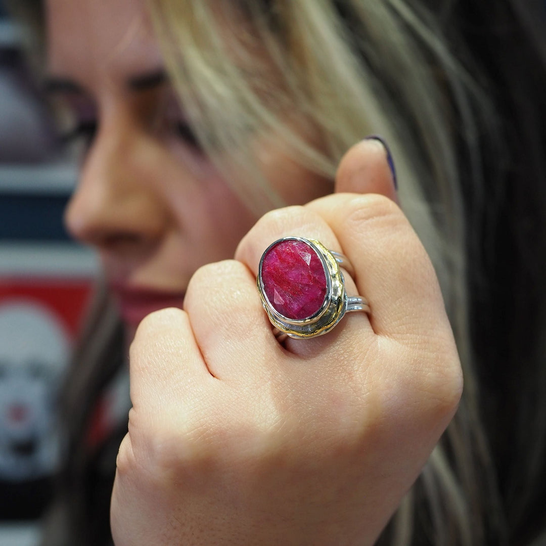 Close-up of a person holding up a hand to display a large, oval red gemstone set in a silver band with a gold accent. The Magnolia Ring in silver gold & various gemstones by Gallardo & Blaine Designs stands out as an exquisite statement ring, with the blurred background focusing attention on the ring and the person's hand.