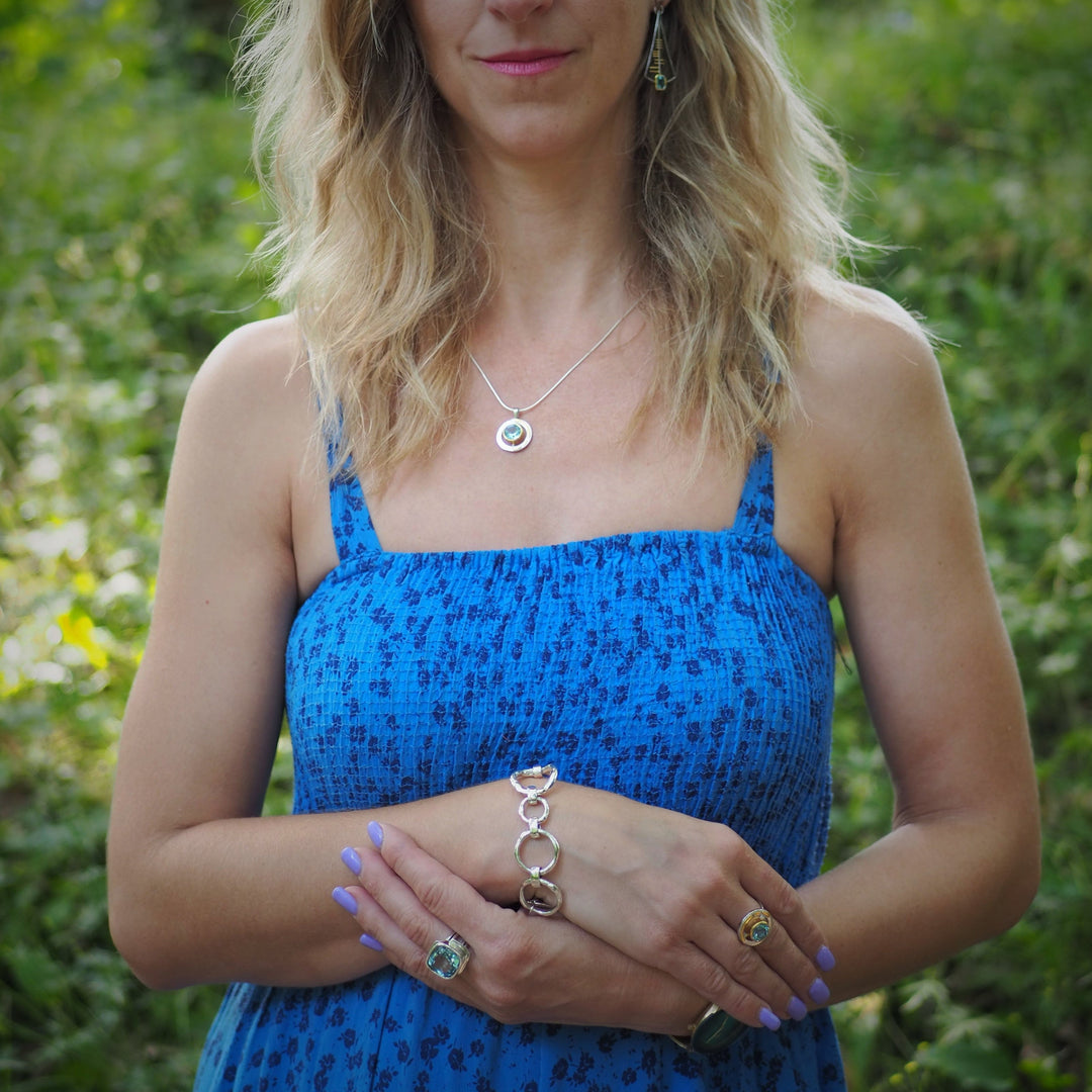 A woman with wavy blonde hair wears a sleeveless blue dress and stands outdoors. She is adorned with jewelry, including earrings, a necklace, and a bracelet. Her hands are positioned in front of her, showcasing an exquisite Art Deco Ring in Silver Gold & various gemstones by Gallardo & Blaine Designs and a sterling silver bracelet.