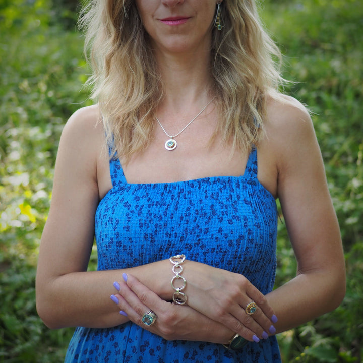 A woman with wavy blonde hair wears a sleeveless blue dress and stands outdoors. She is adorned with jewelry, including earrings, a necklace, and a bracelet. Her hands are positioned in front of her, showcasing an exquisite Art Deco Ring in Silver Gold & various gemstones by Gallardo & Blaine Designs and a sterling silver bracelet.