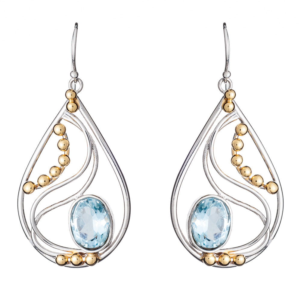 These Phoenix Earrings in various gemstones by Gallardo & Blaine Designs are a pair of elegant drop earrings featuring a combination of silver and gold elements. With a silver frame adorned with intricate swirls and small gold beads, each earring showcases a large, oval light blue gemstone at the center.