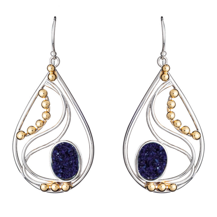 A pair of elegant drop earrings featuring silver-tone metal with curving lines and golden bead accents. The center of each Phoenix Earrings in various gemstones from Gallardo & Blaine Designs displays a deep blue oval gemstone, adding a striking contrast to the metallic elements.
