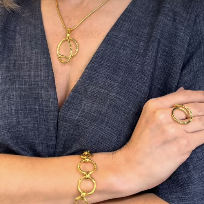 Wearing a deep blue textured V-neck top, the person elegantly displays Gallardo & Blaine Designs’ Sea Honey Ring—featuring interlocking circles—alongside matching gold jewelry: a necklace and bracelet, with arms gracefully crossed.