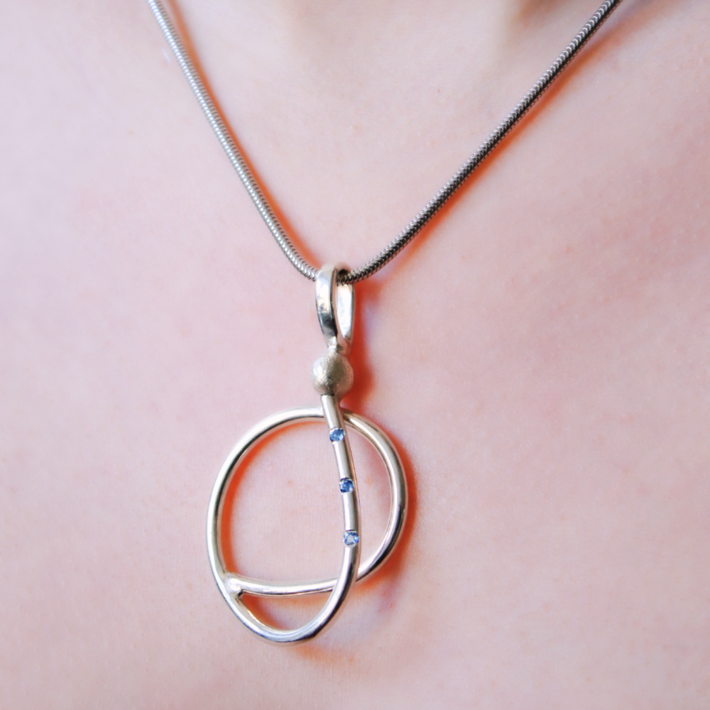 A close-up of Gallardo & Blaine Designs’ Sea Honey Necklace: a sterling silver piece with two intersecting oval loops, accented with small gemstones on one side, and worn on a thin, smooth chain around the neck.