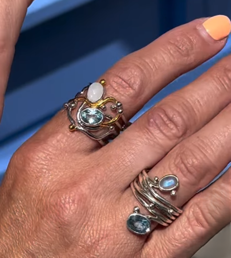 A hand with a peach manicure wears three Gallardo & Blaine Designs sterling silver rings, including the Seaweed Ring in various gemstones, each featuring artistic designs against a softly blurred, blue-toned background.