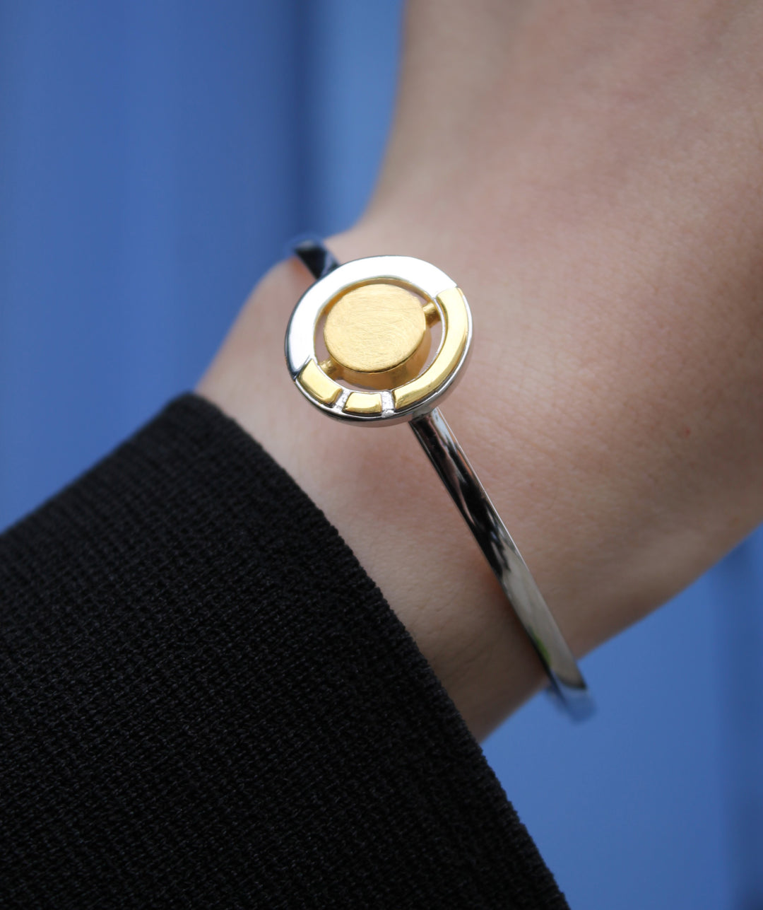A close-up shows a wrist wearing the Urú Cuff Silver & Gold bracelet by Gallardo & Blaine Designs, featuring a sleek sterling silver band with a round gold accent. The person is dressed in black against a solid blue background.