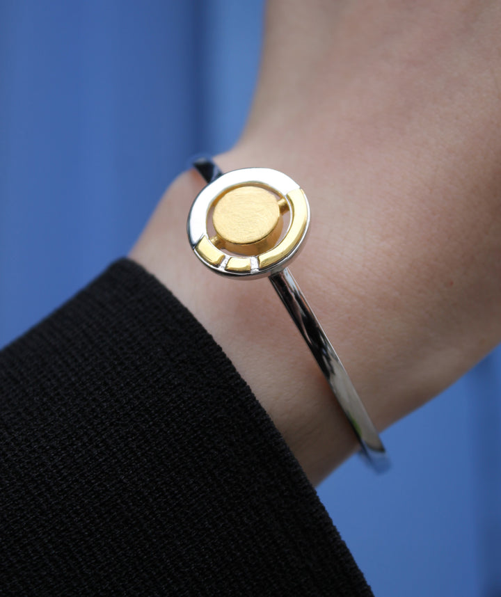 A close-up shows a wrist wearing the Urú Cuff Silver & Gold bracelet by Gallardo & Blaine Designs, featuring a sleek sterling silver band with a round gold accent. The person is dressed in black against a solid blue background.