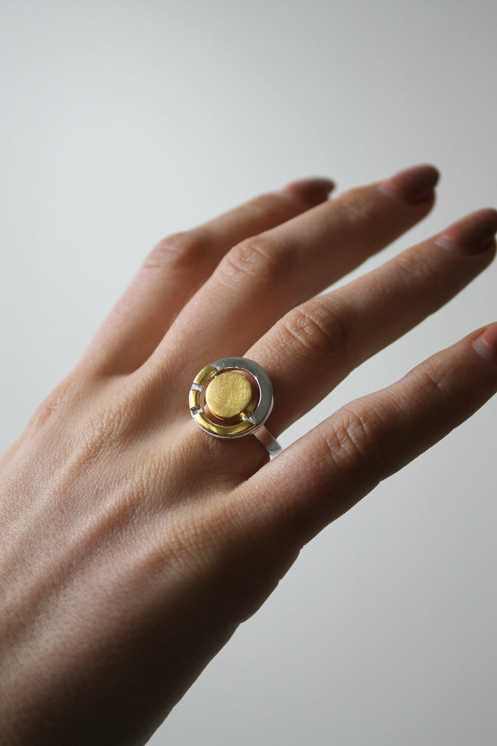 A hand with neatly manicured nails wears the Gallardo & Blaine Designs Urú Ring, a silver Celtic piece with a large, round, gold-colored centerpiece, set against a neutral light background.