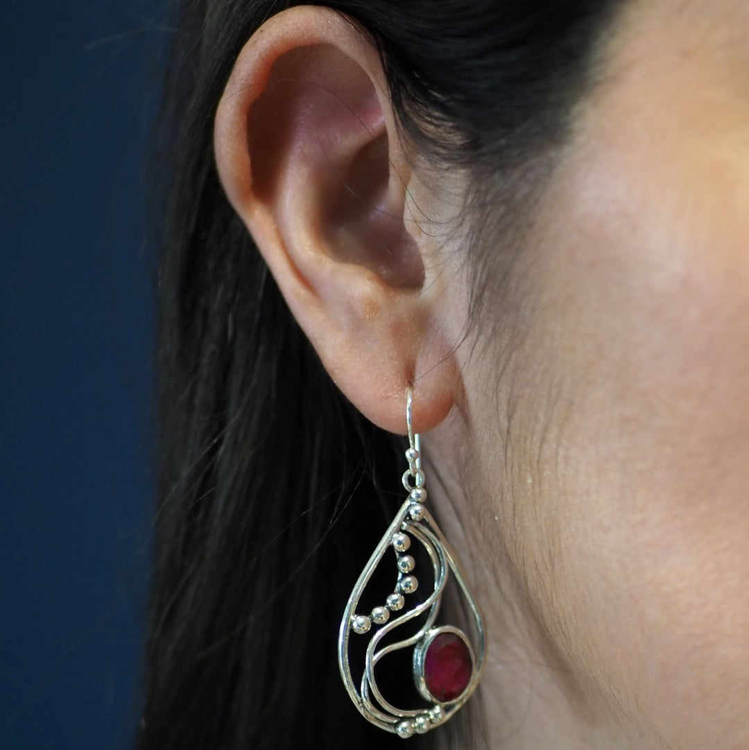 Close-up of a woman's ear wearing a large, intricately designed Phoenix Earrings in various gemstones. The earring is silver with a teardrop shape, adorned with small spherical elements and a striking red oval gemstone near the bottom. The dark background highlights the earring's elegant details. This exquisite piece is by Gallardo & Blaine Designs.