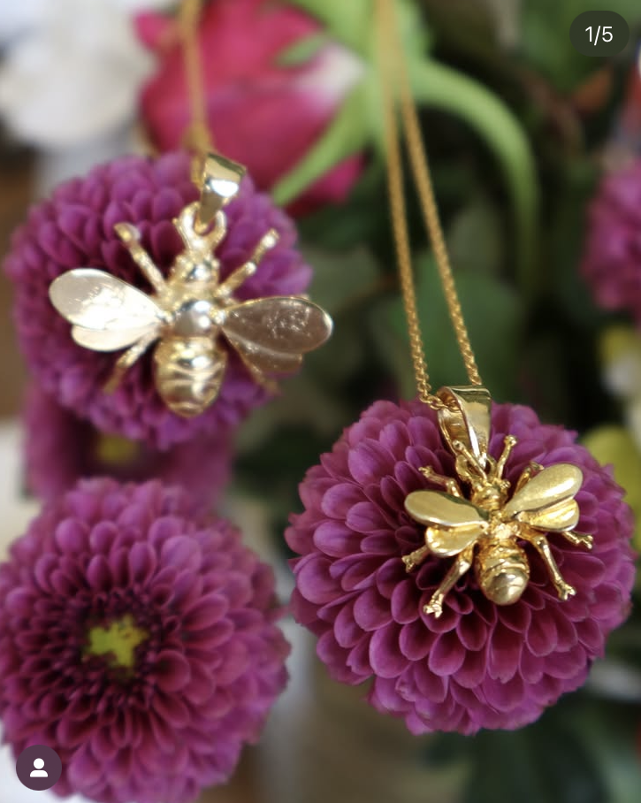 The Gallardo & Blaine Designs Wildlife Pendant - Large Bee With Chain rests atop vivid purple flowers with green centers, highlighting the brand’s elegant, nature-inspired jewelry amidst a dreamy floral backdrop.