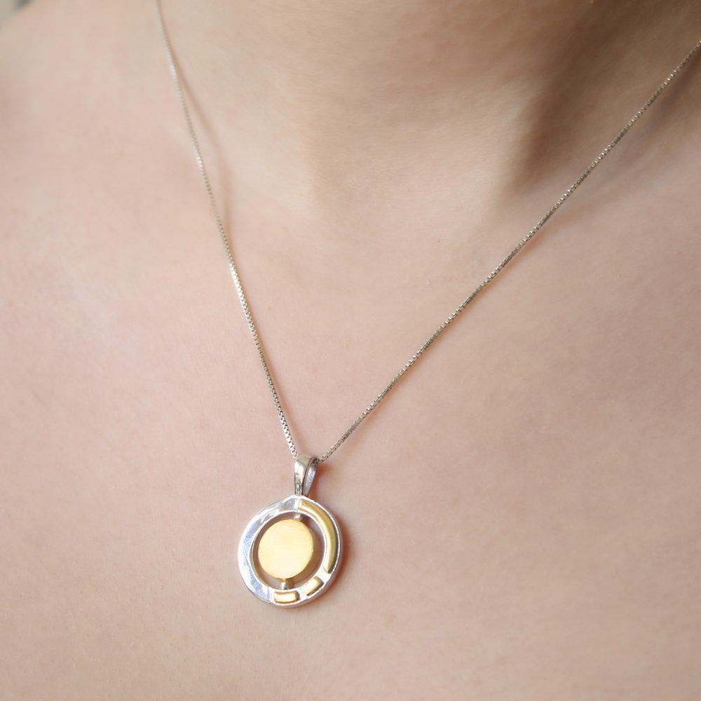 A close-up of a person’s neck wearing the Urú Small Pendant & Chain by Gallardo & Blaine Designs, featuring a modern, minimalist round pendant that elegantly blends sterling silver and gold tones.