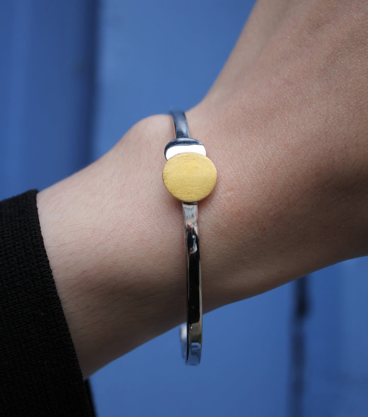 The Archeol Cuff in Sterling Silver & Gold Vermeil by Gallardo & Blaine Designs adorns a wrist, its round gold accent shining against a blurred blue background.