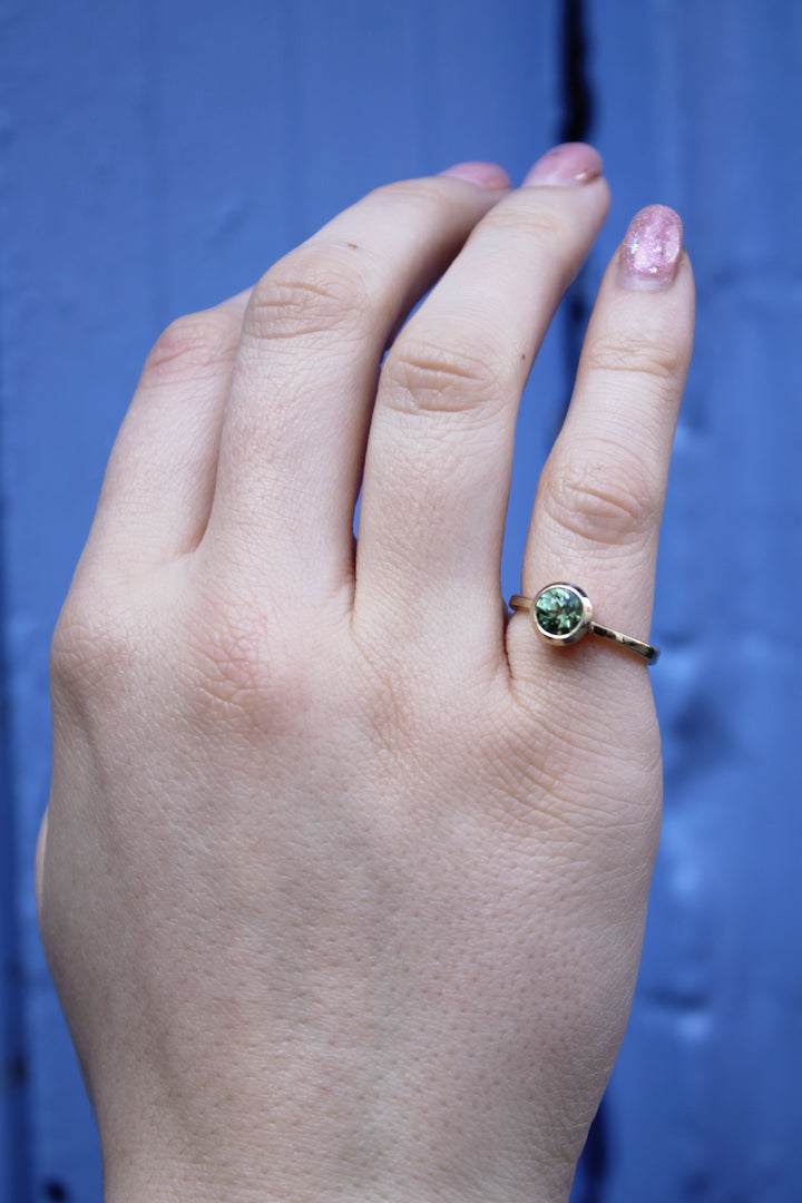 A hand with light skin and pink glittery nail polish wears the Madeleine Viridian Dawn 9ct yellow gold ring, featuring a round gemstone, held up against a blue background.
