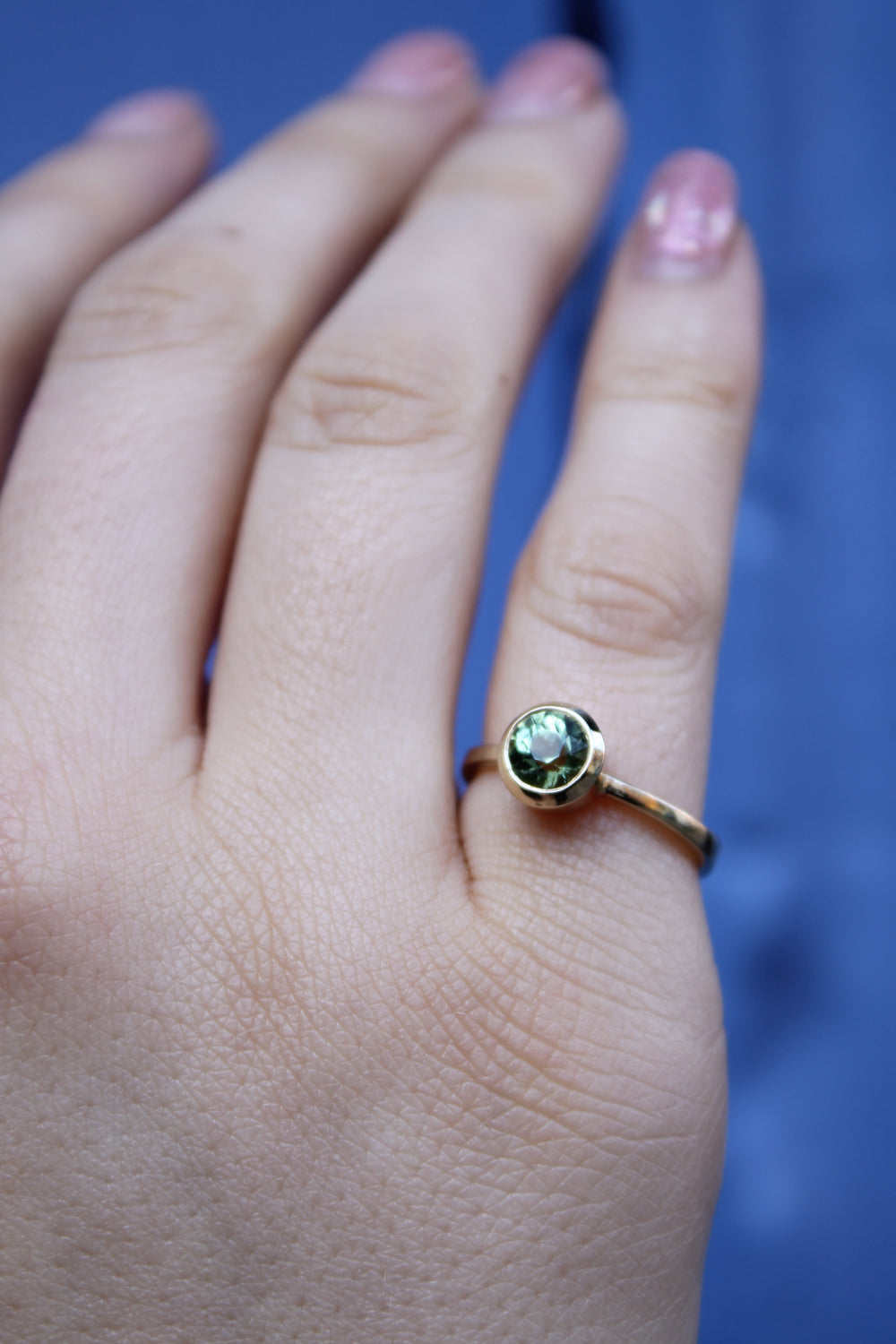 A hand with light skin wears the Madeleine Viridian Dawn 9ct yellow gold ring against a blurry blue background. The short nails are polished in soft light pink.