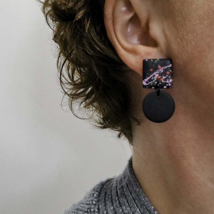 Close-up of a person wearing Culu Design's Hiro Black Splash Earrings—handmade polymer clay with a colorful, abstract square top and black circular pendant—standing out against their short, curly brown hair.