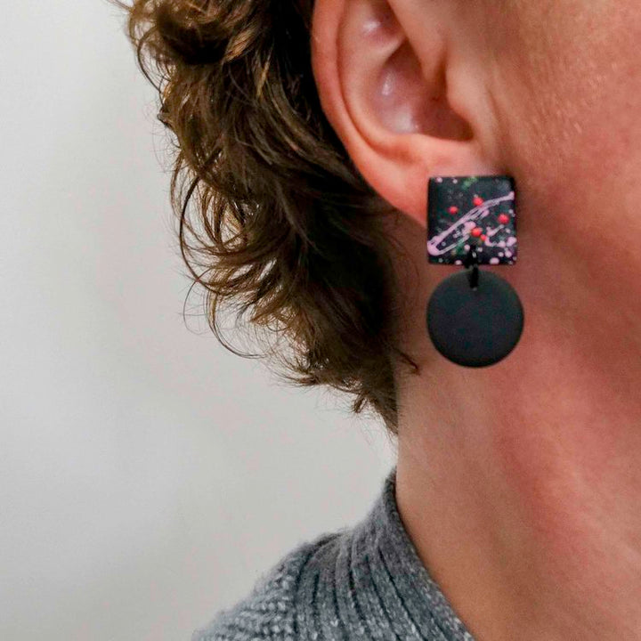 Close-up of a person wearing Culu Design's Hiro Black Splash Earrings—handmade polymer clay with a colorful, abstract square top and black circular pendant—standing out against their short, curly brown hair.