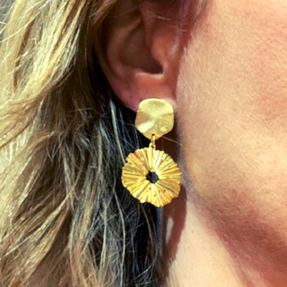 Gold drop, statement earrings. The top of the earring  is a small, irregularly shaped metallic piece that attaches to the earlobe. It has a slightly curved surface with a subtle, organic texture, resembling a petal.   Hanging from the top piece is a circular pendant with a hole in the centre, giving it a donut-like shape. The surface of this pendant features radiating ridges, resembling the folds of a flower or the texture of a sunburst. The ridges create depth and reflect light dynamically.