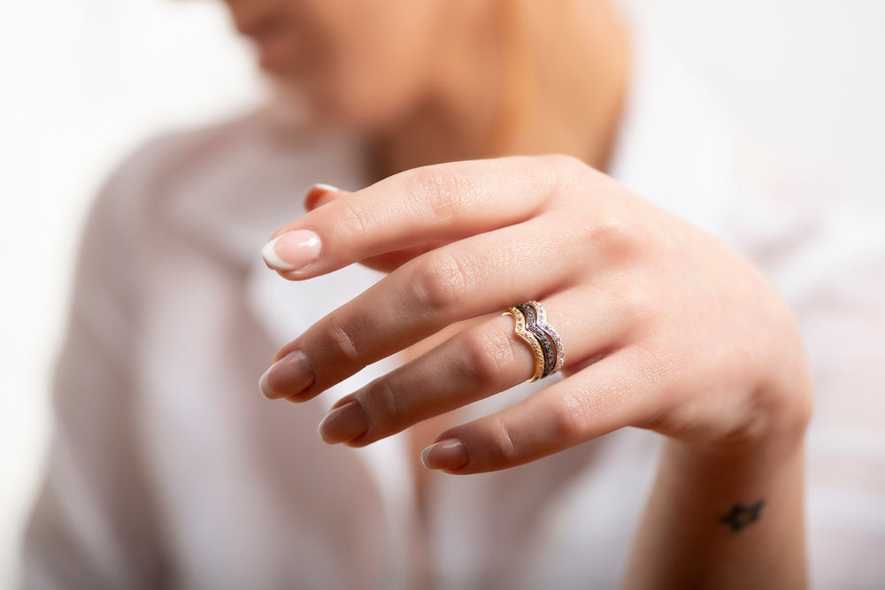 A woman's manicured hand wears two Georgia Charal Imagination Wishbone Rings with colorful zircon on her ring finger. She is dressed in a white shirt, with a small star tattoo on her wrist, and her face is out of focus.