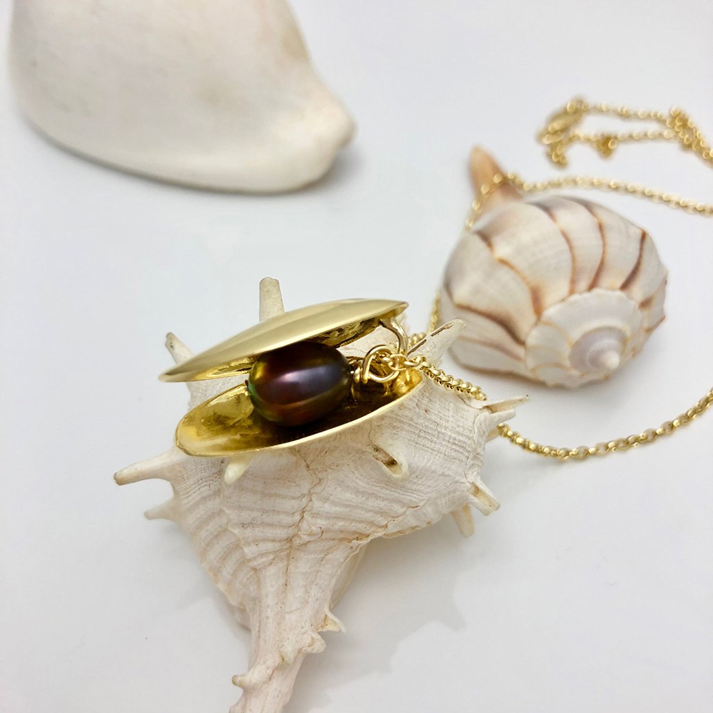 An Open Clam Shell Necklace With Pearl, crafted by Jennifer Kinnear with a gold-plated silver finish, features a dark freshwater pearl nestled inside its clam-shaped pendant. The elegant piece rests on a white spiked seashell. In the background, another seashell and part of a gold chain accentuate the marine-themed jewelry against a plain white surface.