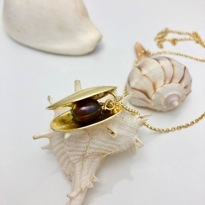 An Open Clam Shell Necklace With Pearl, crafted by Jennifer Kinnear with a gold-plated silver finish, features a dark freshwater pearl nestled inside its clam-shaped pendant. The elegant piece rests on a white spiked seashell. In the background, another seashell and part of a gold chain accentuate the marine-themed jewelry against a plain white surface.