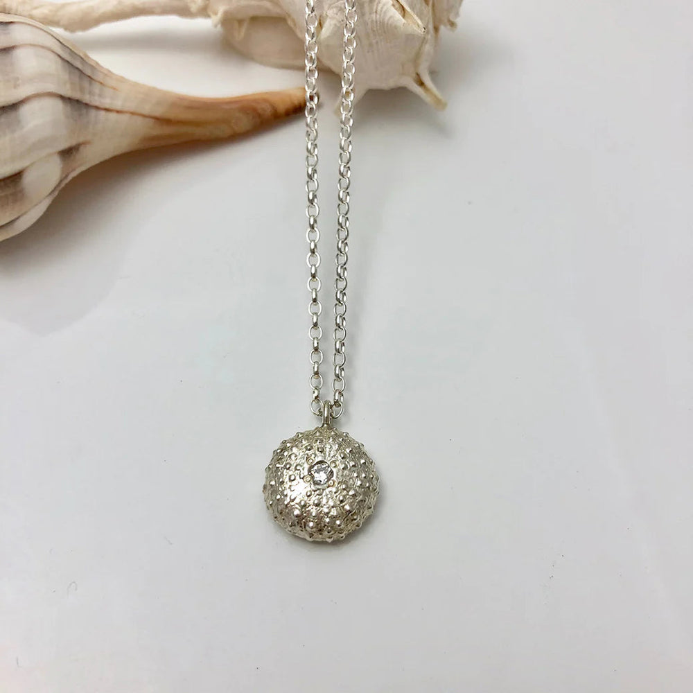 The Jennifer Kinnear Sea Urchin Necklace with Stone in silver or gold features a delicate chain and central gem, shown on a white surface with a seashell in the background.