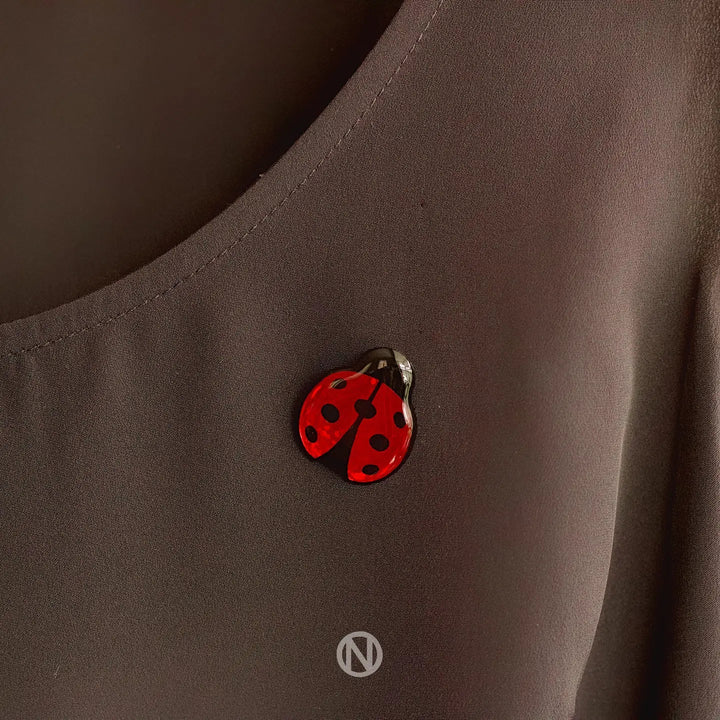 A small, hand-painted Ladybird Pin Brooch with black spots is pinned on a dark-colored fabric, possibly a shirt. The whimsical touch of the ladybug contrasts beautifully with the plain background. A logo featuring an "N" within a circle is visible at the bottom of the image, adding to its distinct charm from Naoi.