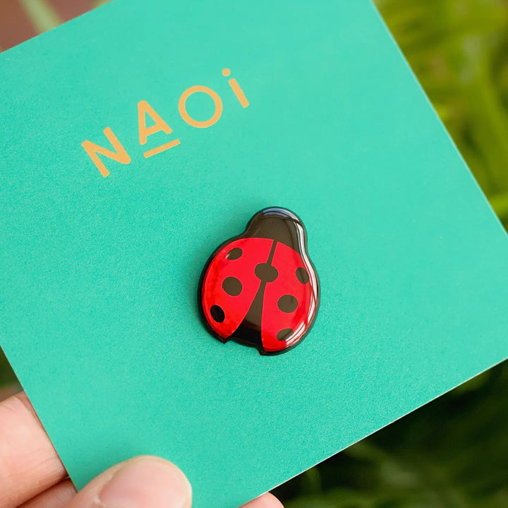 Close-up of a hand holding an enamel pin in the shape of a hand-painted Ladybird Pin Brooch against a turquoise card with the text "Naoi" printed on it. The background appears to be a blurred natural setting.