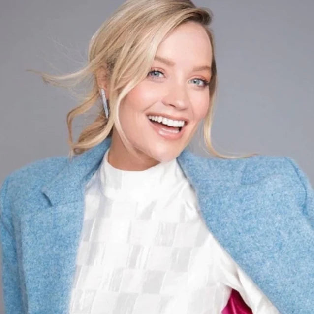 A blonde woman with loose waves smiles at the camera, wearing a light blue blazer and white textured top, showcasing Caroline Stokesberry-lee Raw Kyanite Shard Earrings against a plain light gray background.