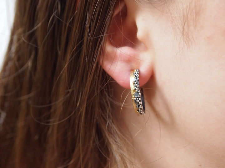 Close-up of a person's ear with straight brown hair, wearing a small gold Etrusc Hoop Earring by MarCucurella, adorned with black and white stones and delicate Etruscan granulation.