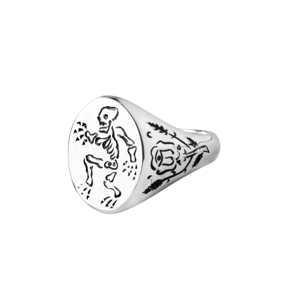 The Sterling Silver Memento Mori Signet Ring by Cosmic Boulevard showcases an engraved skeleton on the round face and a thorned rose etched along the side.