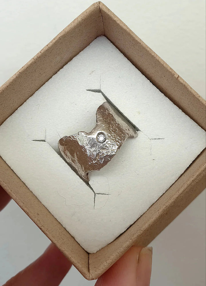 The Morrigan's Faerie Tale Ring by Caroline Stokesberry-lee is a rough, irregularly shaped silver ring with a small round cubic zirconia. It comes in a square cardboard box with white cushioning, held from the bottom corner.