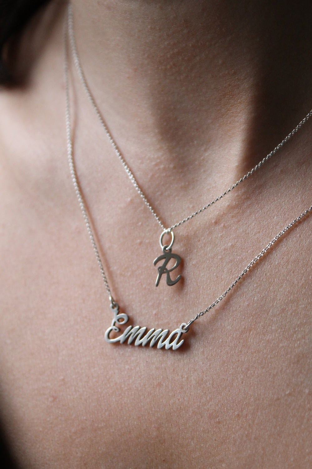 A close-up of a person's neck wearing two silver Personalised Charms necklaces: a Name Necklace with "Emmi" in cursive and an initial pendant shaped like the letter "R.