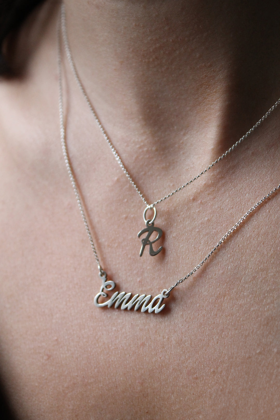 A close-up of a person's neck wearing two silver Personalised Charms necklaces: a Name Necklace with "Emmi" in cursive and an initial pendant shaped like the letter "R.