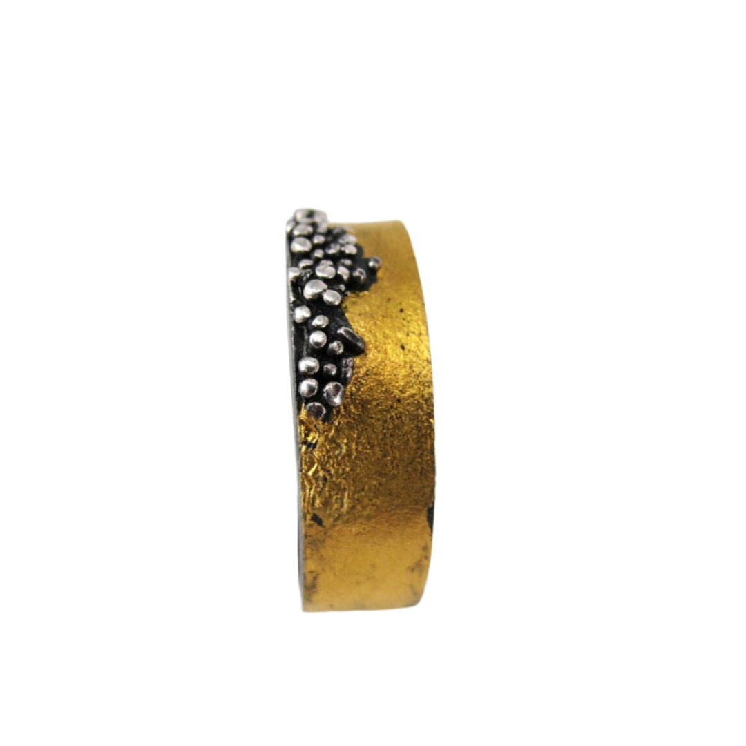 The MarCucurella Narrow Etrusc Ring is gold-colored with textured granulation, featuring a cluster of small oxidized silver beads arranged organically along the top edge, displayed from the side against a white background.