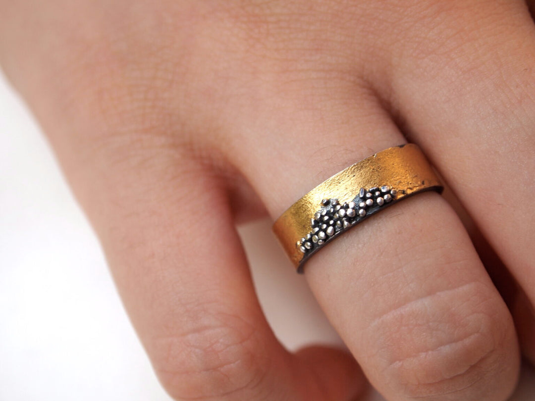 A close-up of a hand wearing the MarCucurella Narrow Etrusc Ring, crafted in 24 kt yellow gold and oxidized silver, featuring textured bands and a cluster of small raised beads in an Etruscan-inspired design.