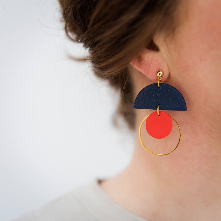 Close-up of a person wearing Olivia Midnight Earrings by Culu Design—handmade polymer clay earrings with a dark blue semicircle, red circle, and gold hoop. Hair is loosely tied back against a plain background.