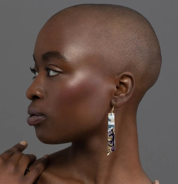 A person with a shaved head and glowing skin gazes left, wearing Rothlú's Panthea Batik Textile Earrings in Dove/Gold/Aubergine. Their hand rests on their shoulder against a smooth gray background.