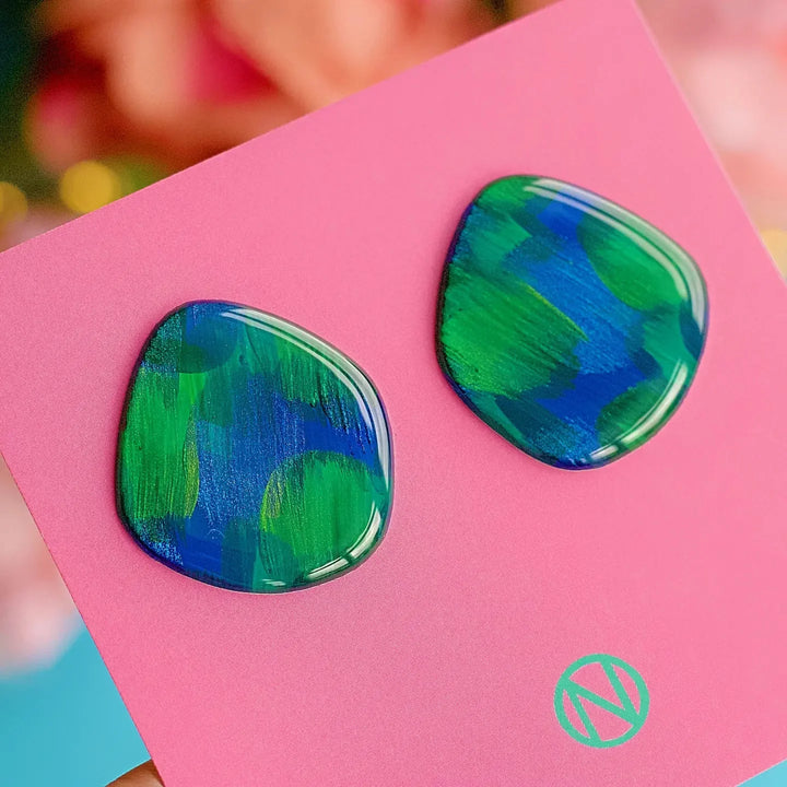 Peacock Vibes Stud Earrings by Naoi: hand-painted blue and green brushstrokes on irregular shapes, displayed on a pink card with a circular logo. Made in Ireland from lightweight paper.