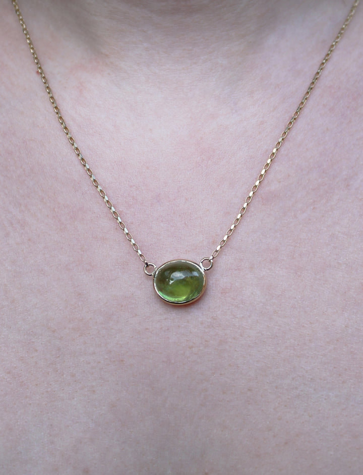A close-up of the Madeleine Whispering Moss 9ct Yellow Gold Necklace, featuring a round green gemstone pendant, worn around the neck.