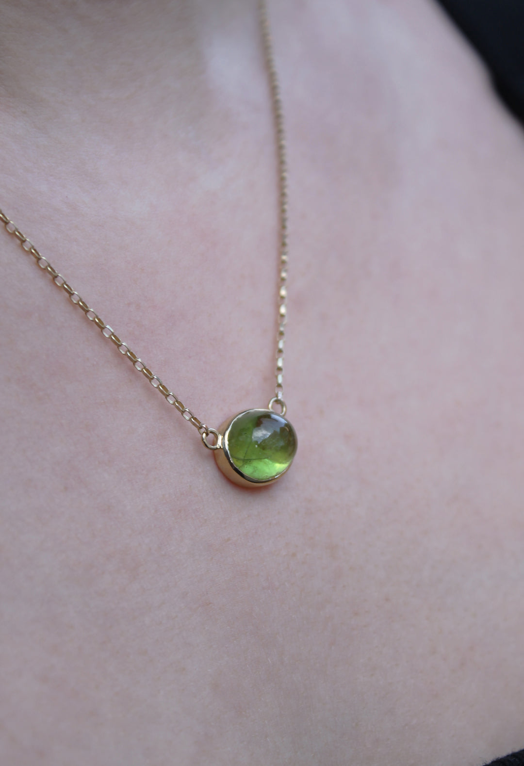 A close-up of the Madeleine Whispering Moss 9ct Yellow Gold Necklace with an oval cabochon peridot pendant resting on a person’s skin.