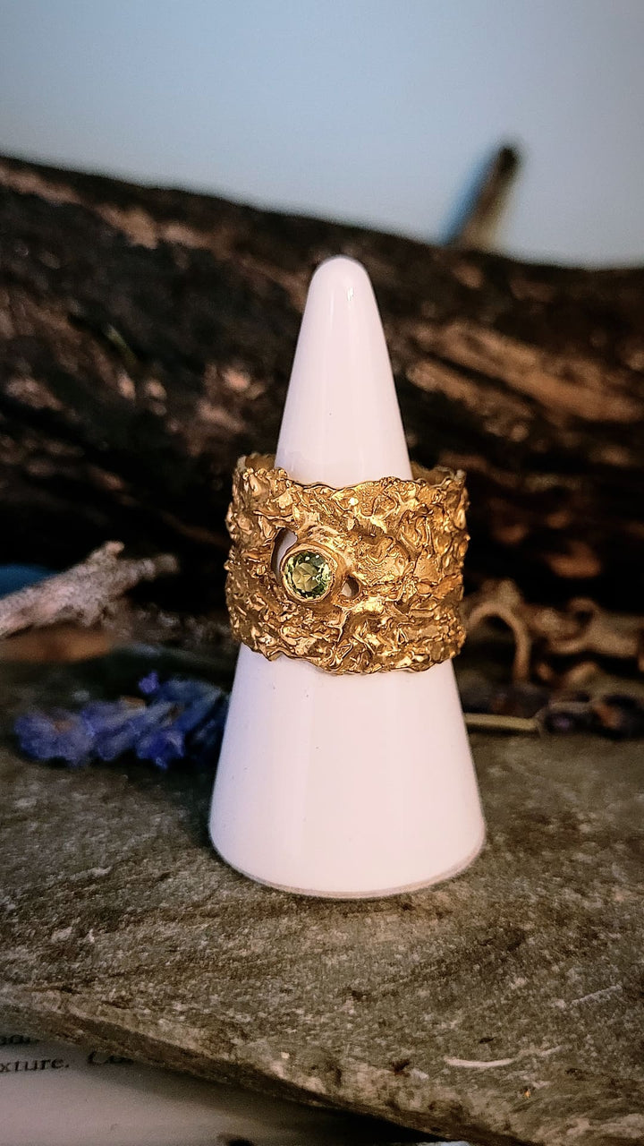 The 'That Place Beyond' Wide Ring by Caroline Stokesberry-lee features a green gemstone in 18ct gold plate, shown upright on a white cone stand with a rustic wooden background.